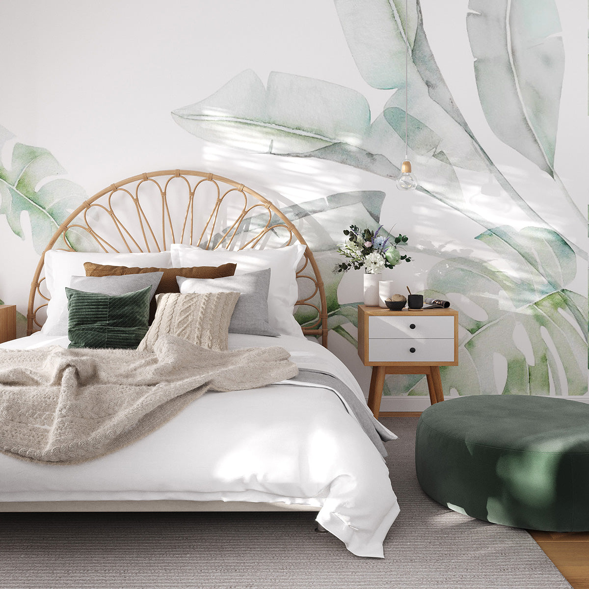 Soft green leaf wallpaper mural with oversized watercolour banana and monstera leaves on a white background behind a rattan bed and a green velvet pouffe